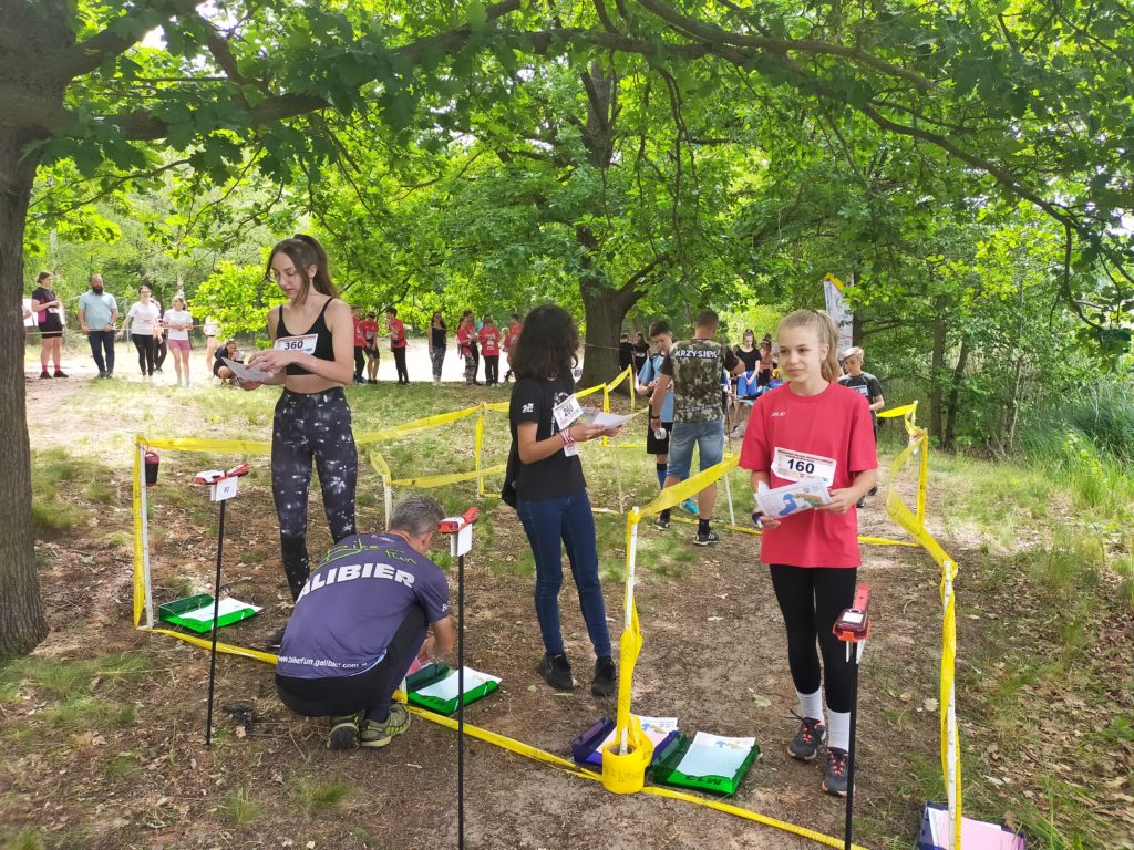 Zdjęcie przedstawia trzy zawodniczki stojące na linii startu z mapami do biegu w rękach w zadrzewionym terenie